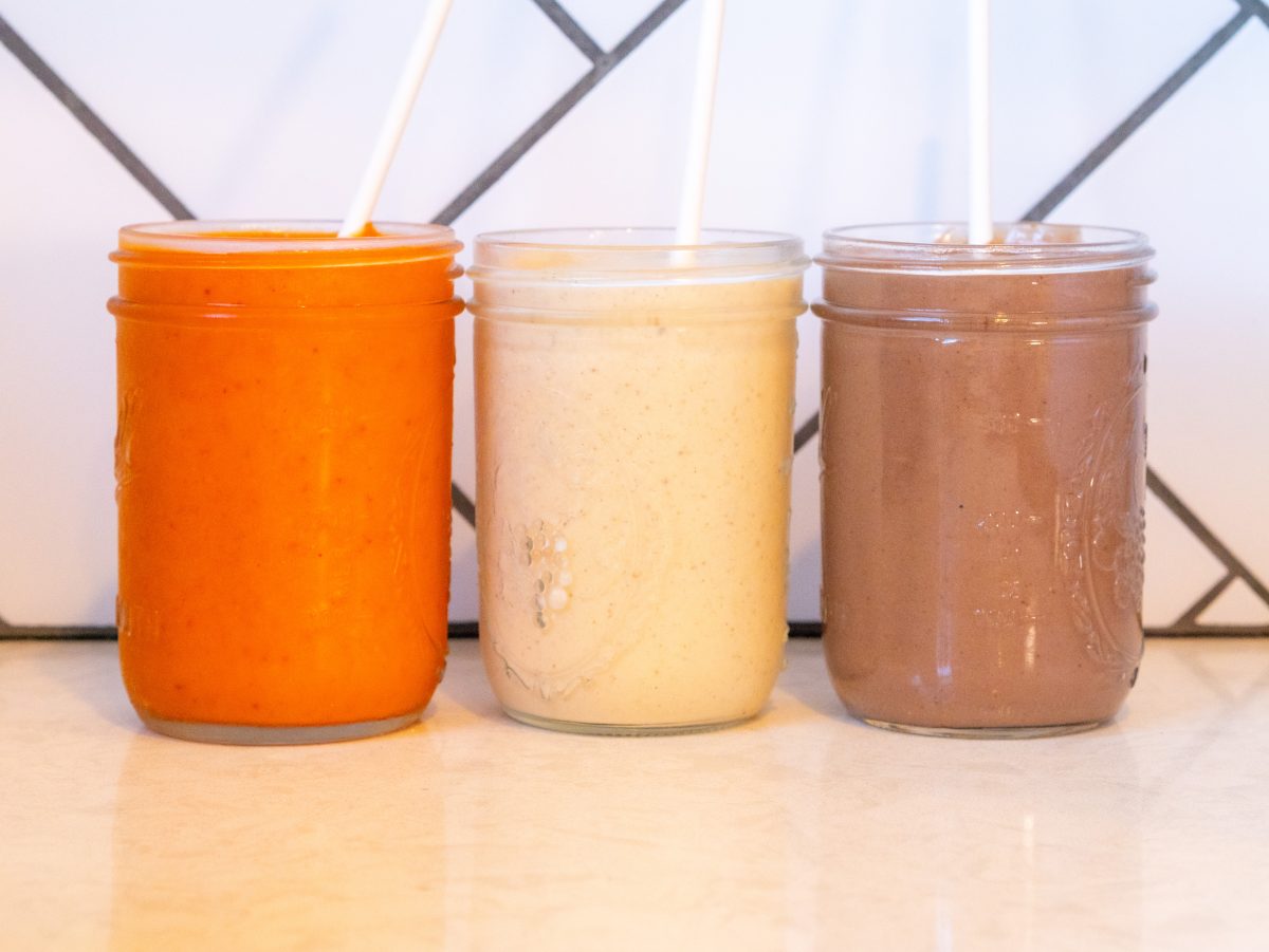 Three colorful dairy-free smoothies made with frozen fruit served in glass jars including peaches and cream, chocolate peanut butter, and tropical fruit.