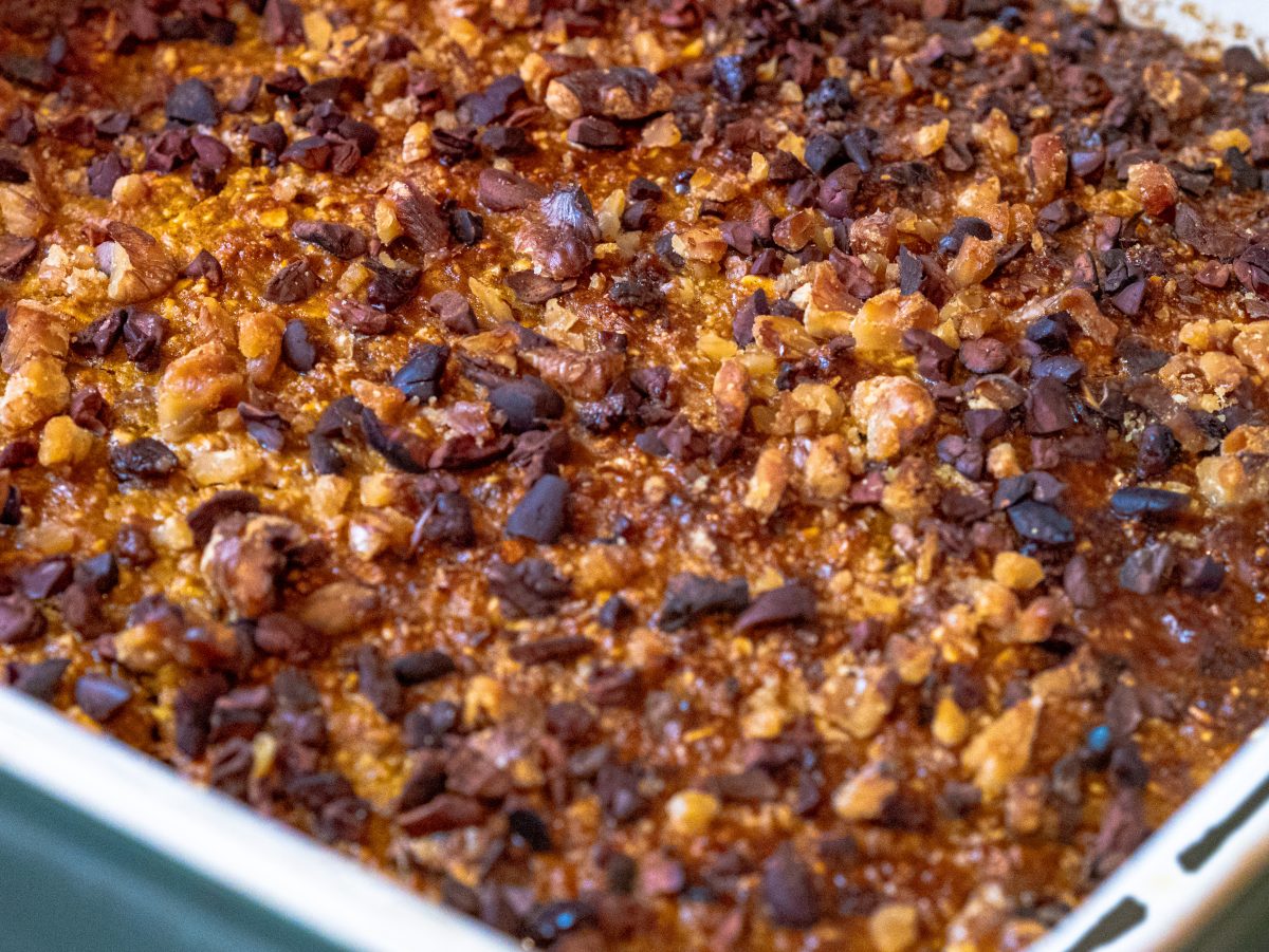 Warm pumpkin spiced baked oats in a square baking dish, featuring puffed golden edges and a topping of chopped walnuts and dark cocoa nibs.